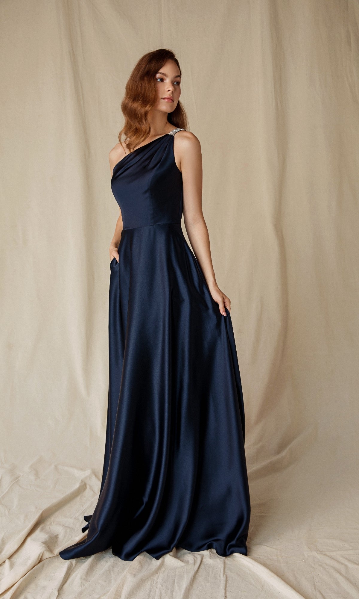 A woman stands on a neutral fabric backdrop wearing the Eureka 9025, a floor-length, dark blue one-shoulder satin formal dress with an asymmetrical neckline, holding the skirt with one hand.